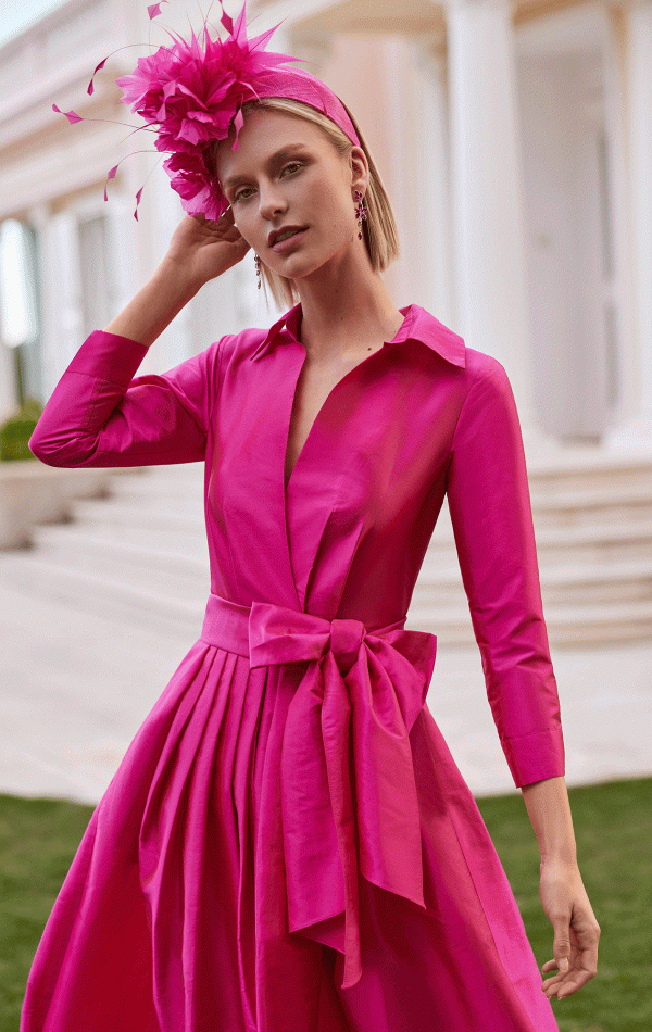 A person in a bright pink dress, by the Rosa Clara Couture Club - 9GM8, with a matching headpiece stands elegantly outside a building with grand white columns.