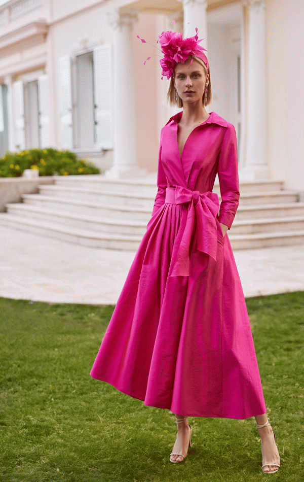 Wearing the Rosa Clara Couture Club - 9GM8, a member dazzles in vibrant pink with a matching hat, gracefully posed on the grass before a grand white building with columns.