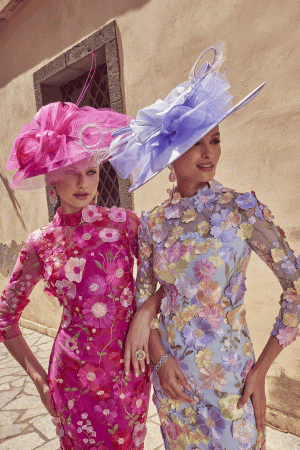 Two women in Veni Infantino 992445 floral dresses and large hats pose elegantly outside a building.