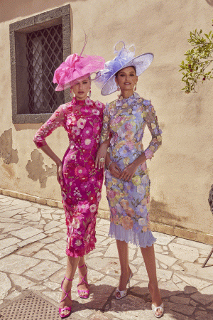 Two women wearing Veni Infantino 992445 floral embroidered dresses and large hats pose elegantly outside a beige building on a sunny day.