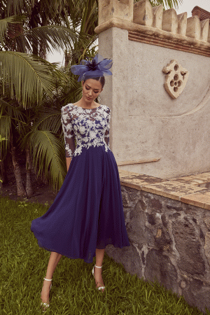 A woman wearing the Veni Infantino - 992450 blue dress with a lace top and fascinator stands beside a stone wall, surrounded by palm leaves.