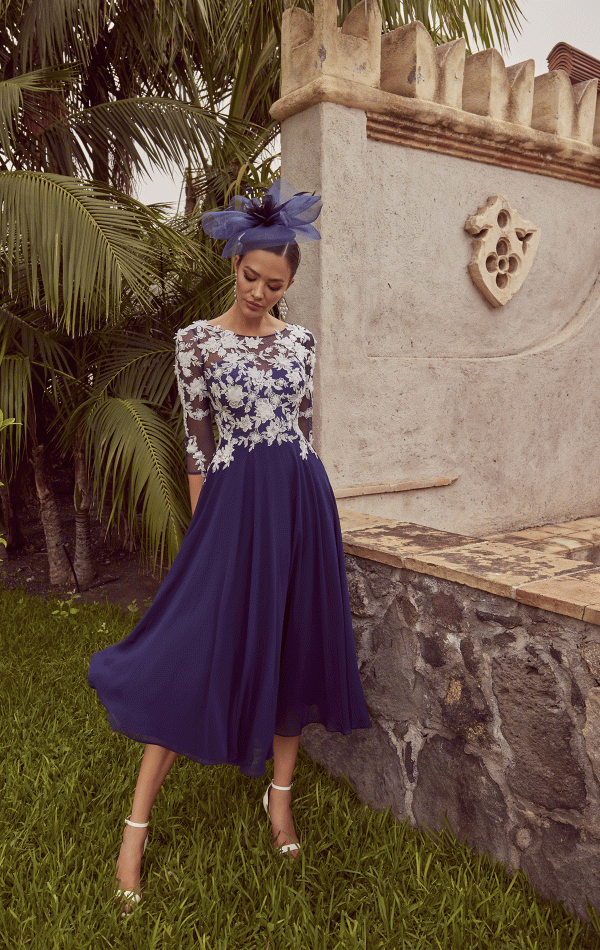 A woman wearing the Veni Infantino - 992450 blue dress with a lace top and fascinator stands beside a stone wall, surrounded by palm leaves.