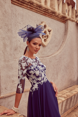 A woman in a Veni Infantino 992450 blue dress with floral lace details and a matching fascinator poses against a decorative wall.