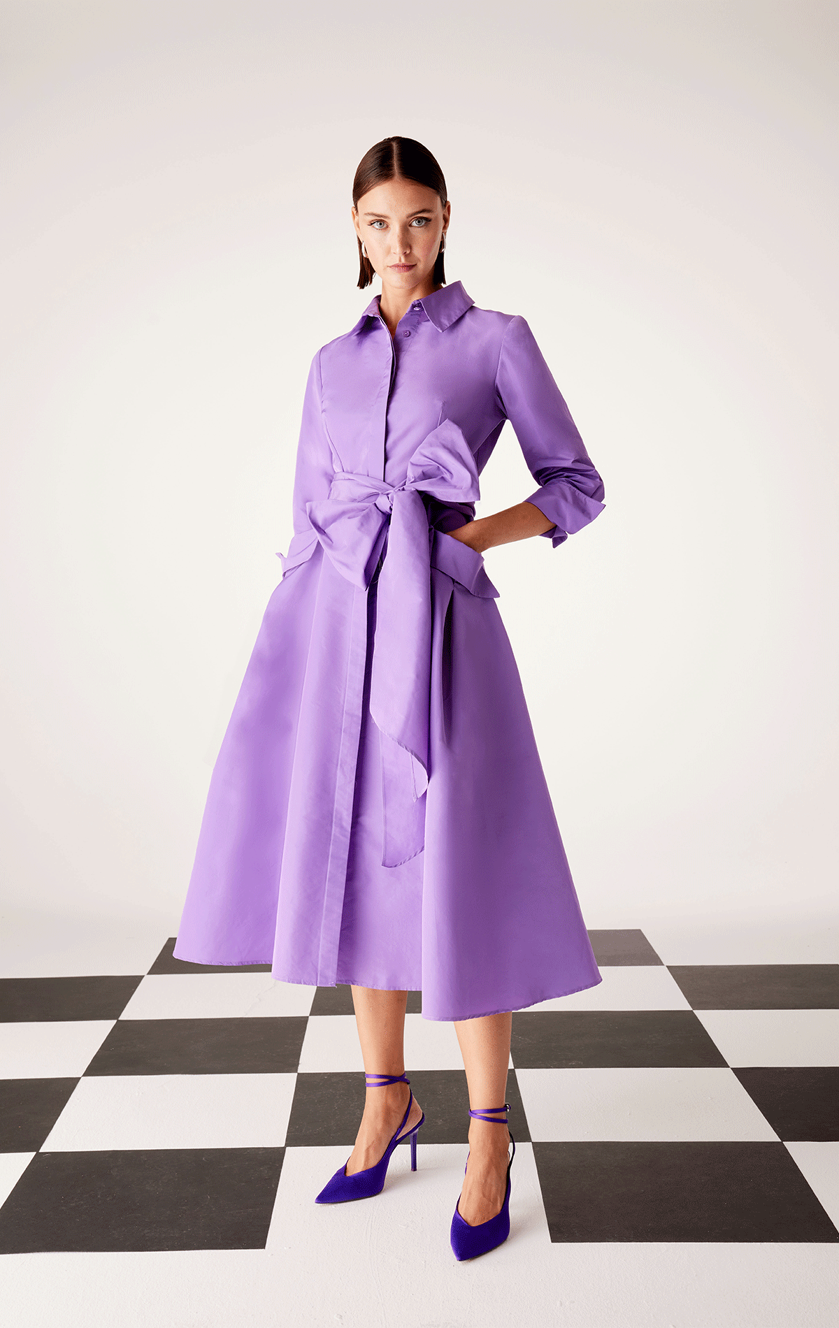A woman in a chic Carla Ruiz - 51547 purple dress and heels stands elegantly on a black and white checkered floor.