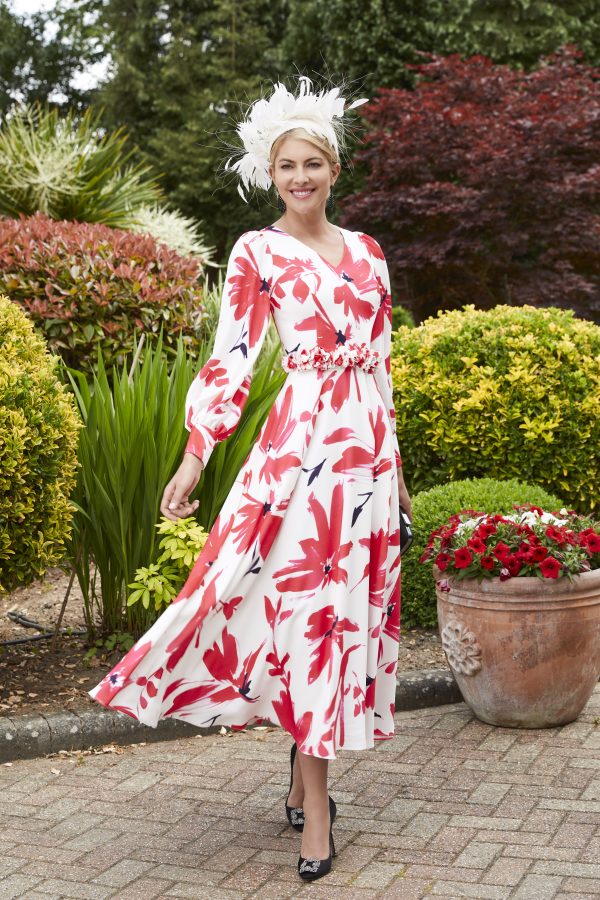 Amid a vibrant garden, a woman gracefully smiles in her Veni Infantino 29679 floral red and white dress, beautifully complemented by a feathered hat.