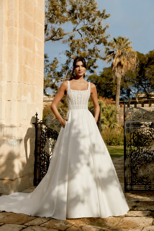 A person in a stylish Sincerity 44493 - Vivienne wedding dress stands by an ornate gate, with a tranquil garden and lush trees creating the perfect backdrop.