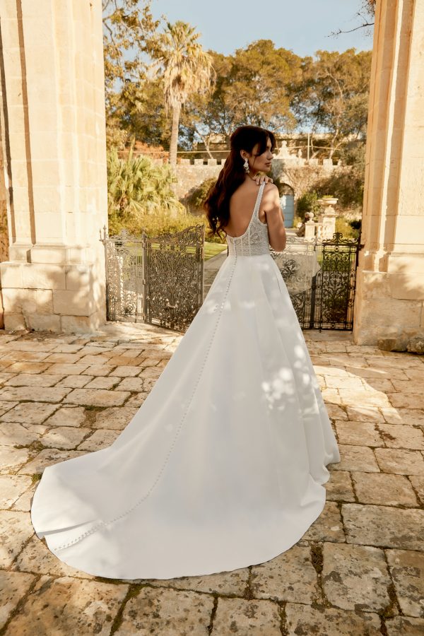 The bride, in her flowing white Sincerity 44493 - Vivienne wedding dress, stands gracefully on the cobblestone, enveloped by majestic stone columns and lush greenery.