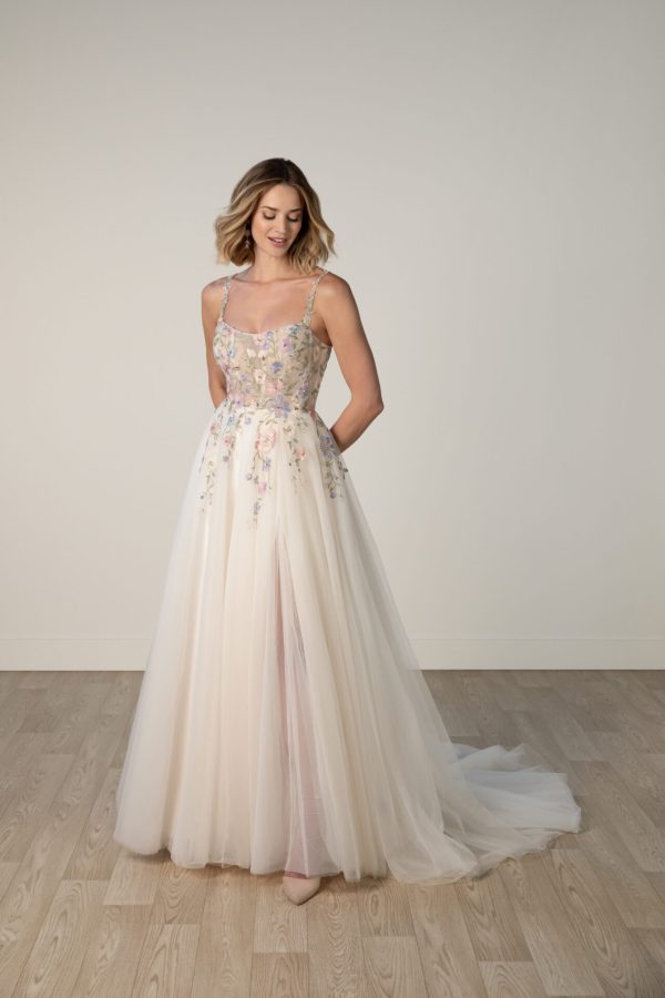 A woman in a Stella York 8063 - Leilani gown stands on a wooden floor, floral embroidery flowing, against a plain backdrop, looking to the side.