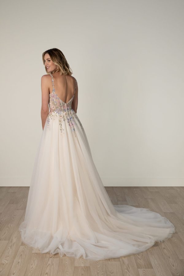 A woman in the Stella York 8063 - Leilani, a long backless white dress with colorful floral details, stands gracefully on a wooden floor, facing away.