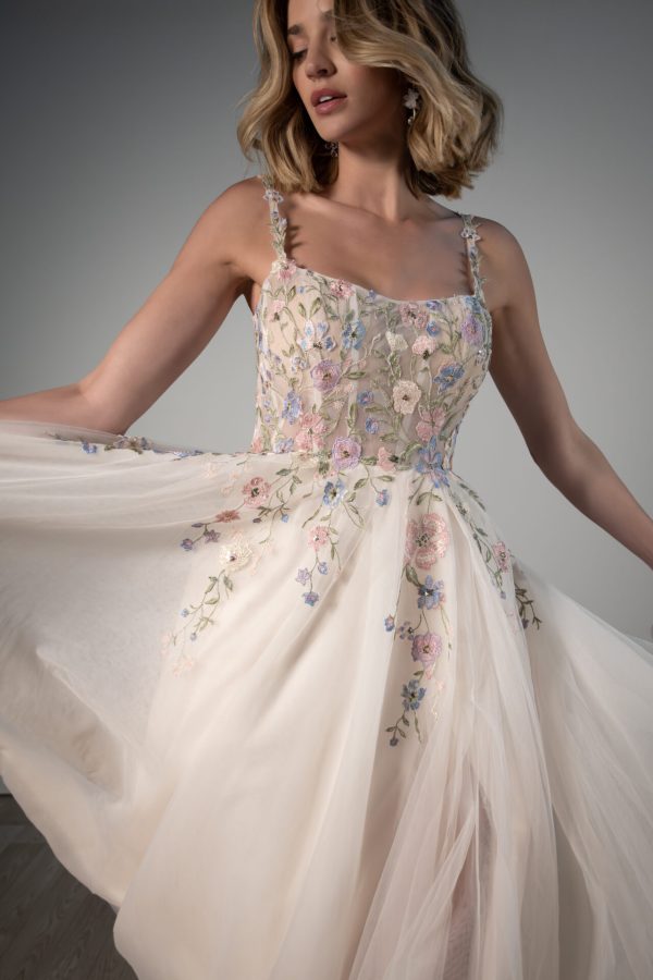 A woman gracefully holds the skirt of a Stella York 8063 - Leilani floral embroidered gown in a softly lit setting.