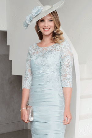 Woman in an Ispirato light blue lace dress and hat smiles indoors, holding a silver clutch (ISJ825).