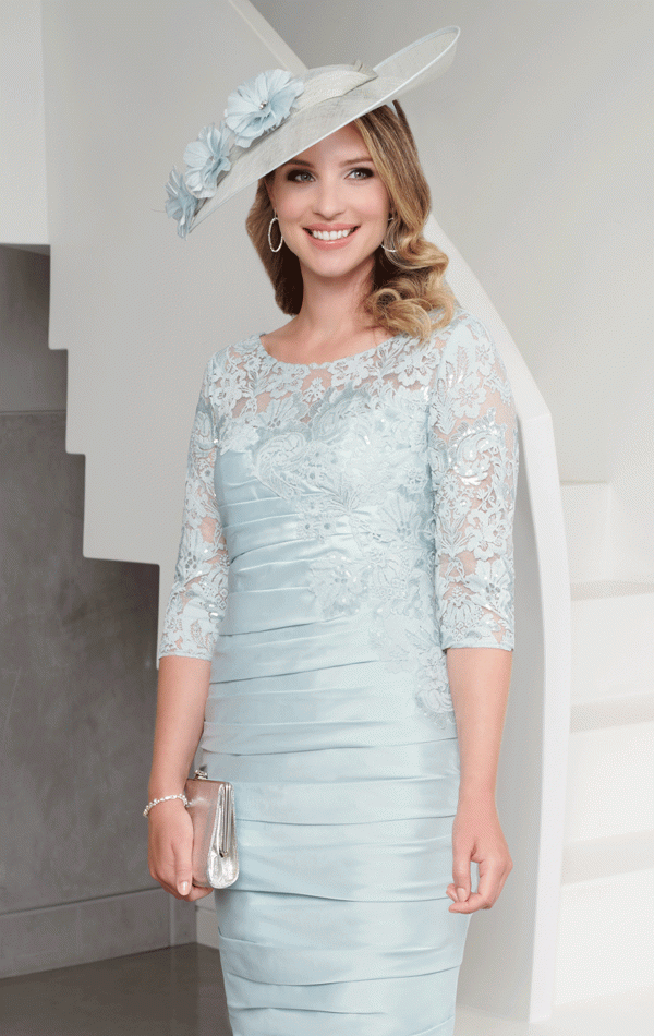 Woman in an Ispirato light blue lace dress and hat smiles indoors, holding a silver clutch (ISJ825).