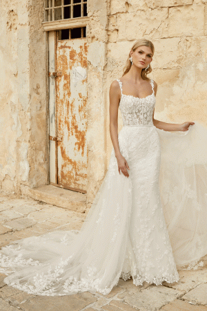 Bride in a Justin Alexander 44482 - Marylise lace wedding dress with train, standing by an aged stone wall and rusty door.