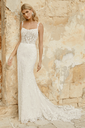 A woman in an elegant Justin Alexander 44482 - Marylise lace wedding dress leans against a textured, weathered wall.
