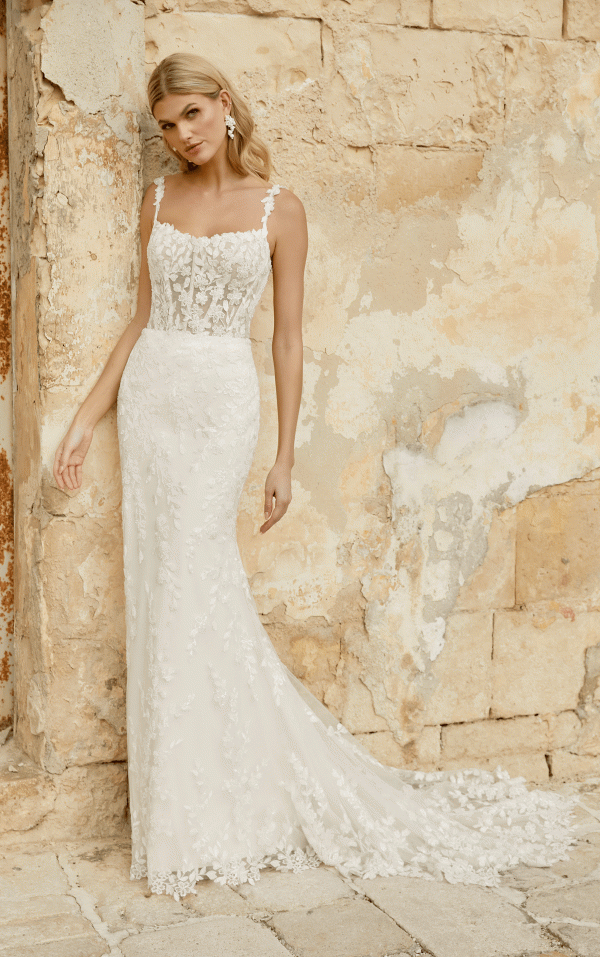 A woman in an elegant Justin Alexander 44482 - Marylise lace wedding dress leans against a textured, weathered wall.