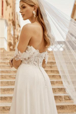The bride, elegant in a Justin Alexander 44485 - Estelle strapless lace gown, stands on stone stairs with her veil gracefully floating as she gazes to the side.