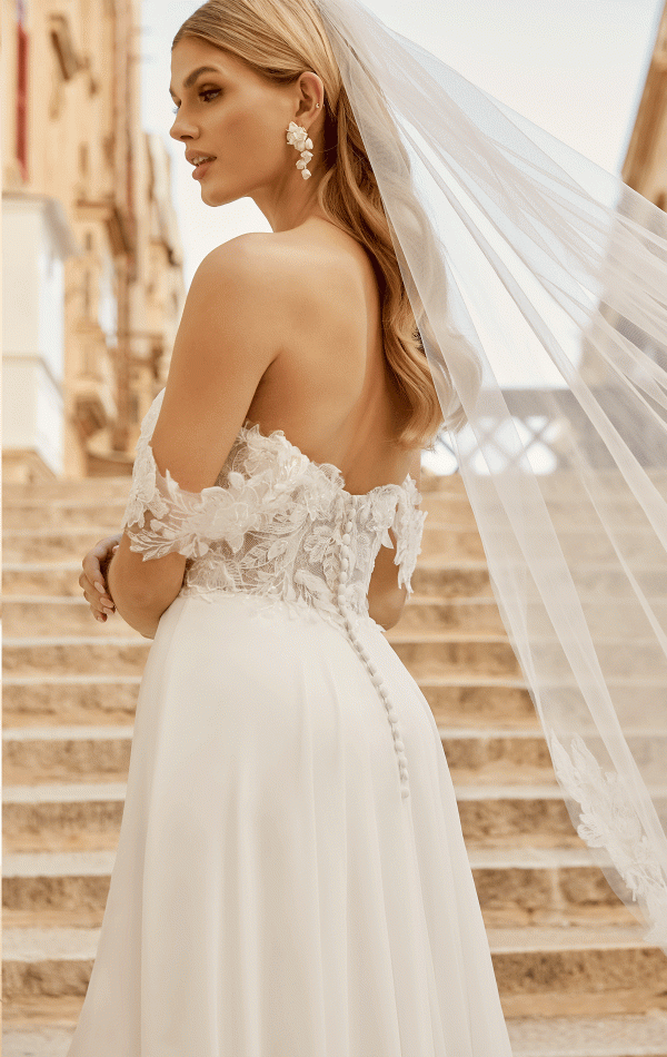 The bride, elegant in a Justin Alexander 44485 - Estelle strapless lace gown, stands on stone stairs with her veil gracefully floating as she gazes to the side.