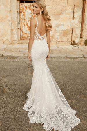 A woman in the Justin Alexander 44482 - Marylise backless lace wedding gown stands gracefully against a rustic wall.
