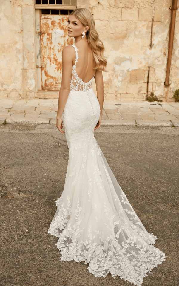 A woman in the Justin Alexander 44482 - Marylise backless lace wedding gown stands gracefully against a rustic wall.