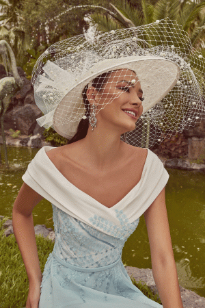 A woman in a Veni Infantino - 992439 blue and white dress, topped with a large white hat, sits happily by a pond surrounded by lush greenery.