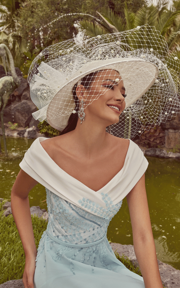 A woman in a Veni Infantino - 992439 blue and white dress, topped with a large white hat, sits happily by a pond surrounded by lush greenery.