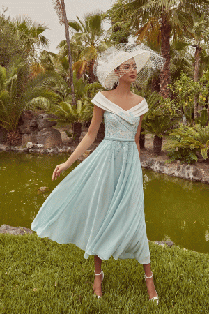 A woman in a Mint green & Ivory Veni Infantino - 992439 dress and wide-brim hat stands by a pond amid timeless fashion.