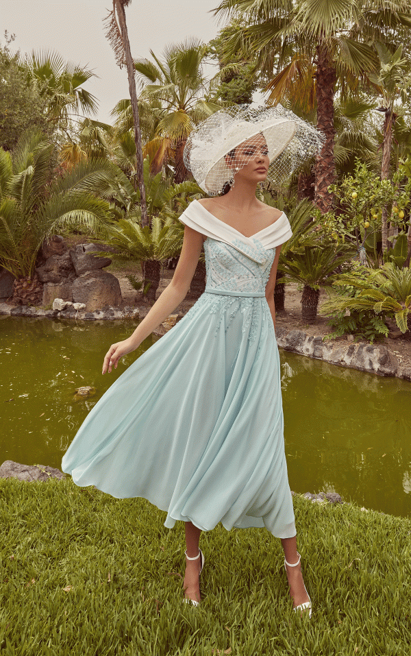 A woman in a Mint green & Ivory Veni Infantino - 992439 dress and wide-brim hat stands by a pond amid timeless fashion.