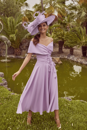 A woman in a Veni Infantino 992446 off-shoulder lavender dress and large hat stands gracefully by the pond, framed by palm trees, embodying timeless fashion.