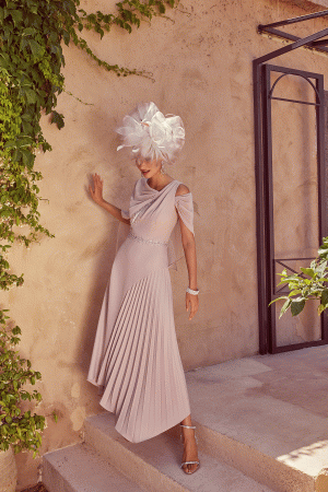 A woman poses elegantly in a flowing Veni Infantino - 992422 pink dress and ornate white hat against a lush green, foliage-covered wall, capturing the essence of timeless beauty.