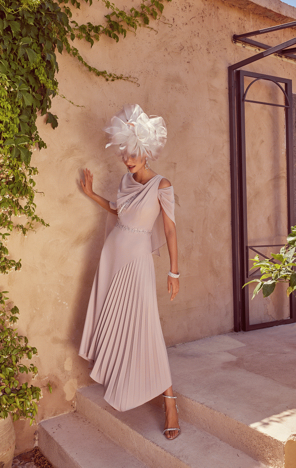 A woman poses elegantly in a flowing Veni Infantino - 992422 pink dress and ornate white hat against a lush green, foliage-covered wall, capturing the essence of timeless beauty.