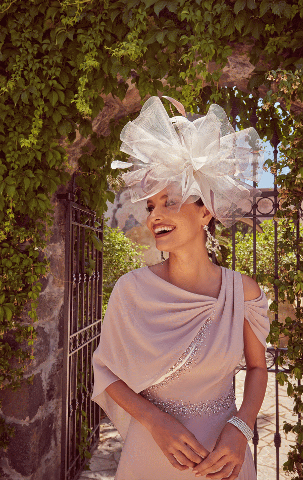 A woman in the Veni Infantino 992422 light pink dress and a large white hat stands gracefully by a vine-covered stone archway.