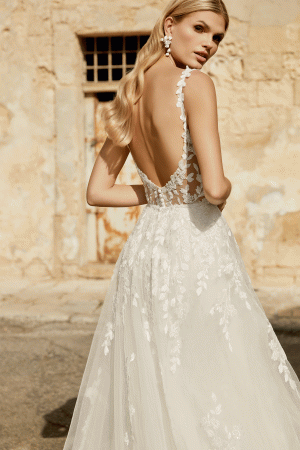 A woman poses outdoors in a backless lace Justin Alexander 44482 - Marylise wedding dress. Her wavy hair cascades, floral earrings glint softly, and she exudes elegance against the textured charm of a rustic wall.