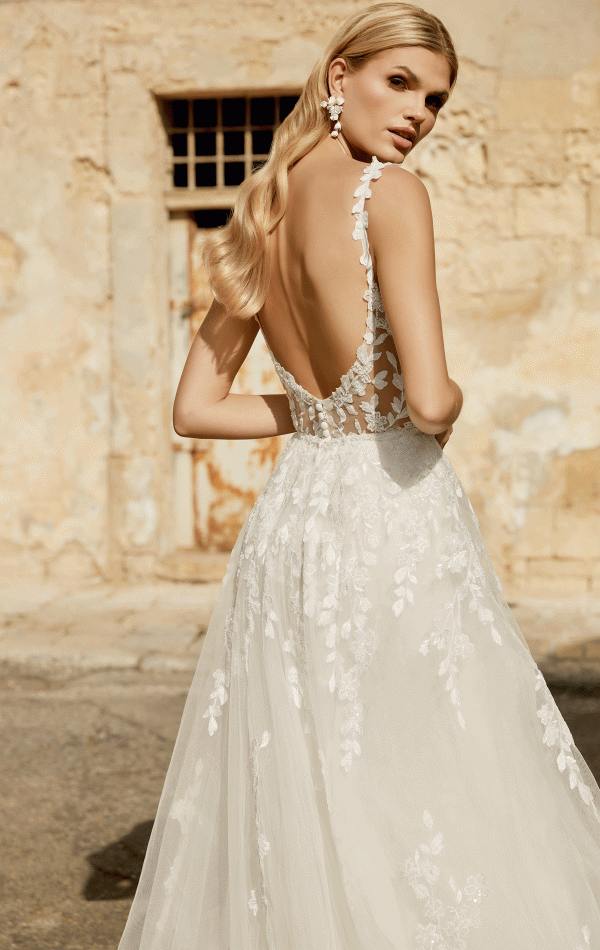 A woman poses outdoors in a backless lace Justin Alexander 44482 - Marylise wedding dress. Her wavy hair cascades, floral earrings glint softly, and she exudes elegance against the textured charm of a rustic wall.