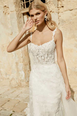 The bride, wearing the Justin Alexander 44482 - Marylise lace dress, stands outdoors against a rustic wall, gently touching her ear.