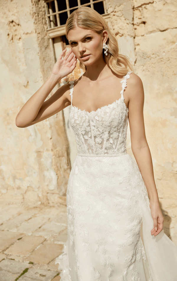 The bride, wearing the Justin Alexander 44482 - Marylise lace dress, stands outdoors against a rustic wall, gently touching her ear.