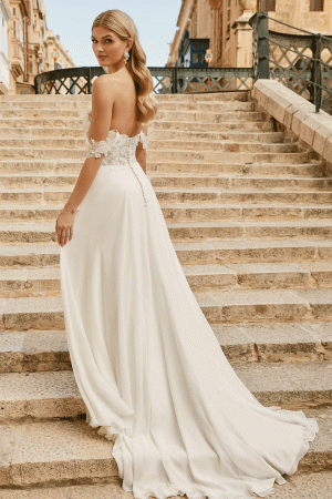 A bride, in a breathtaking Justin Alexander 44485 - Estelle off-shoulder wedding dress, stands on stone steps, gracefully glancing back over her shoulder.