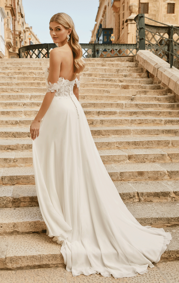 A bride, in a breathtaking Justin Alexander 44485 - Estelle off-shoulder wedding dress, stands on stone steps, gracefully glancing back over her shoulder.