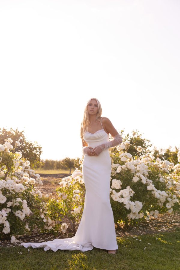 Brooke stands gracefully on the grass in her Stella York 7831 - Brooke wedding dress, surrounded by white flowers with sunlight streaming in the background.