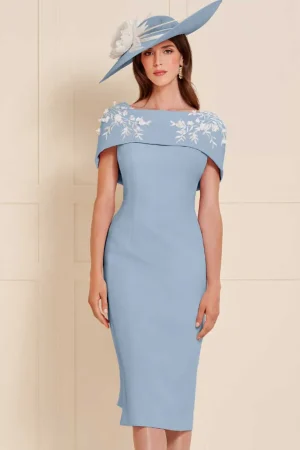 A woman in a light blue John Charles - 66503 dress with a matching hat with floral embroidery stands elegantly against a beige wall.