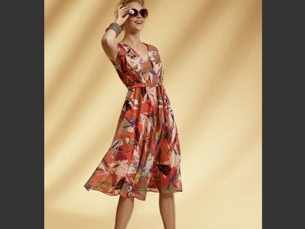 A person in a vibrant floral Arggido - 43630 dress, paired with sunglasses and bracelets, stands against a beige background. Ideal for your next online purchase.