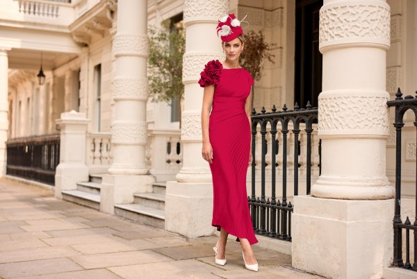 A woman in a red dress and floral hat, embodying the elegance of John Charles 66666, stands by a white-columned building in white heels.