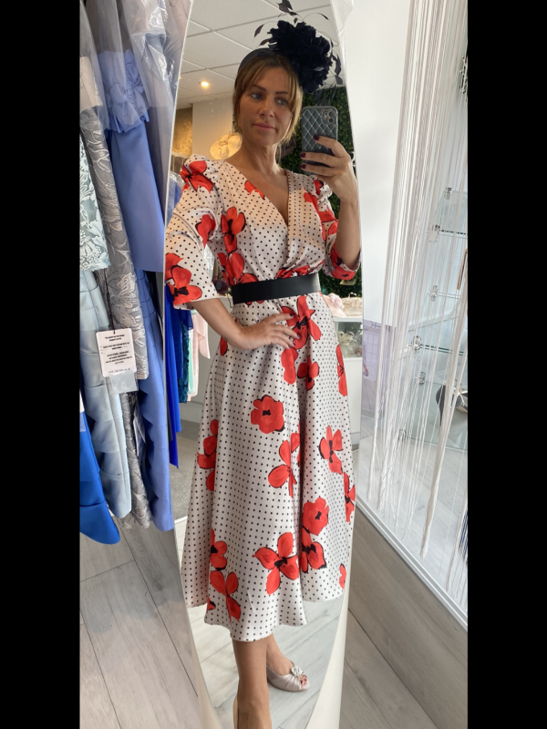 In a chic clothing store, a woman in a floral dress snaps a selfie, flaunting her black belt and elegant fascinator. Her outfit epitomizes Carla Ruiz's stunning designs like the 199624, available for online purchase only.
