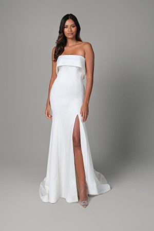 A woman in a strapless white gown with a high slit stands against a gray background, evoking the timeless elegance of Justin Alexander's Isla design.