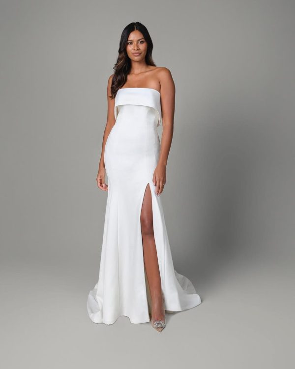 A woman in a strapless white gown with a high slit stands against a gray background, evoking the timeless elegance of Justin Alexander's Isla design.