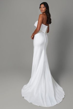 A woman in a strapless, fitted Justin Alexander - Isla gown with a long train stands gracefully, looking over her shoulder, exuding elegance and sophistication.