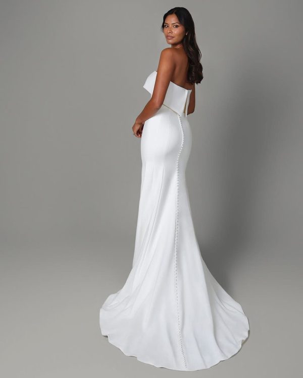 A woman in a strapless, fitted Justin Alexander - Isla gown with a long train stands gracefully, looking over her shoulder, exuding elegance and sophistication.
