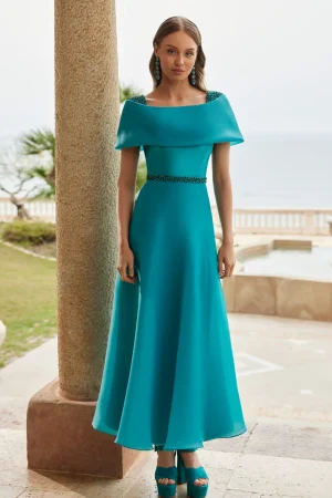 Woman in a turquoise off-shoulder Rosa Clara - 9G199 gown with matching heels, standing outdoors near a seaside terrace.