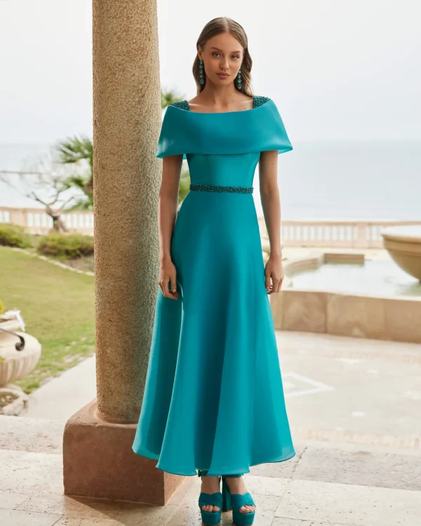 Woman in a turquoise off-shoulder Rosa Clara - 9G199 gown with matching heels, standing outdoors near a seaside terrace.