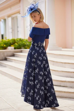 Woman in an off-shoulder navy Rosa Clara - 9G1F3 dress with a white leaf pattern, wearing a blue hat, standing outdoors.