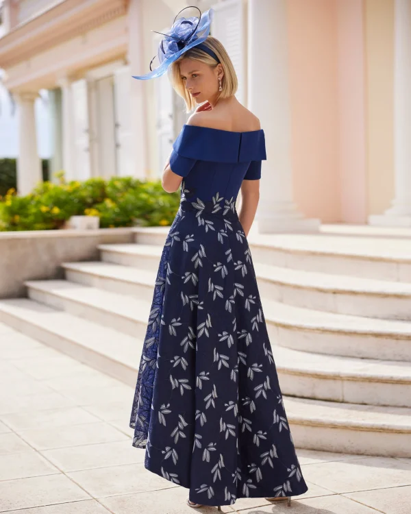 Woman in an off-shoulder navy Rosa Clara - 9G1F3 dress with a white leaf pattern, wearing a blue hat, standing outdoors.